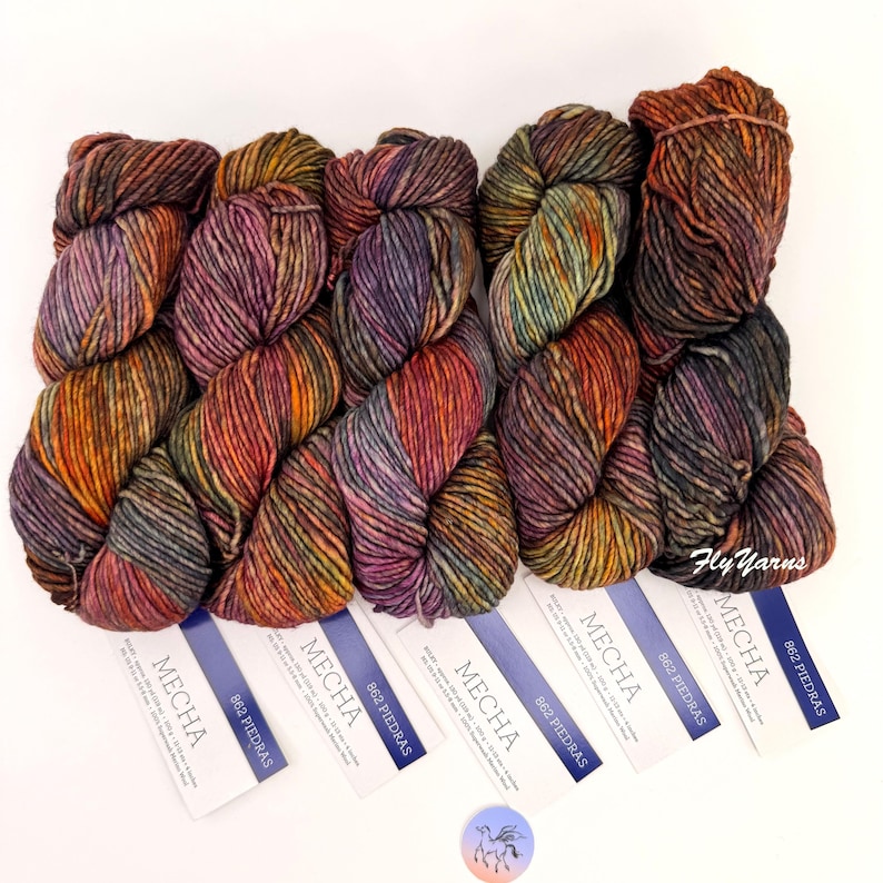 May include: Five skeins of Mecha 862 Piedras yarn, each featuring a variegated blend of purple, orange, green, and brown tones. Each skein is accompanied by a white paper label displaying the brand name Mecha, color code 862 Piedras, and text.