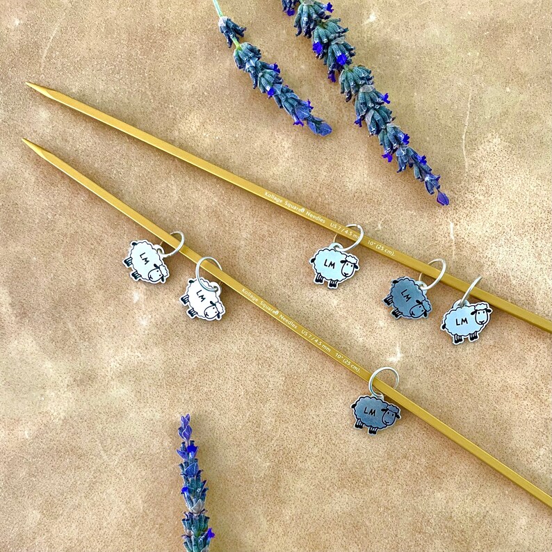 May include: A close-up shot of gold-colored knitting needles with sheep-shaped stitch markers. The stitch markers are white and gray with the initials "LM". Lavender sprigs are arranged around the needles. The needles are 10 inches (25 cm) long.