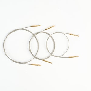 Pu&ograve; includere: Tre ferri circolari con cavi argentati e punte color oro sono disposti su sfondo bianco. Gli aghi variano di dimensioni, creando un aspetto nidificato. L'immagine &egrave; ben illuminata e si concentra sui materiali per l'artigianato.
