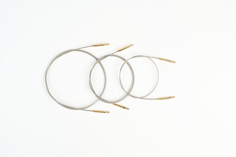 Pu&ograve; includere: Tre ferri circolari con cavi argentati e punte color oro sono disposti su sfondo bianco. Gli aghi variano di dimensioni, creando un aspetto nidificato. L'immagine &egrave; ben illuminata e si concentra sui materiali per l'artigianato.