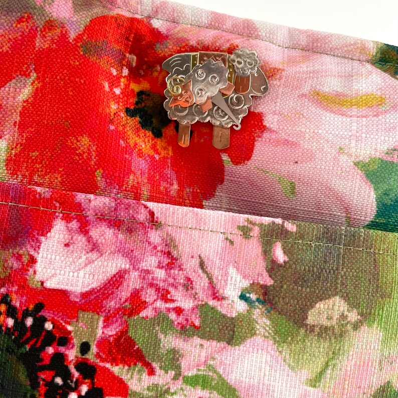Pode incluir: Um broche de metal prateado em forma de ovelha com um design floral sobre um fundo de tecido floral rosa e vermelho.
