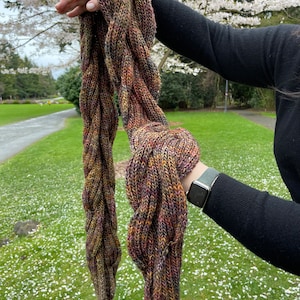 May include: A hand-knitted scarf featuring a thick, braided cable pattern. The yarn is a variegated blend of earthy tones, including deep brown, forest green, burnt orange, and muted purple, creating a textured, multi-colored appearance.