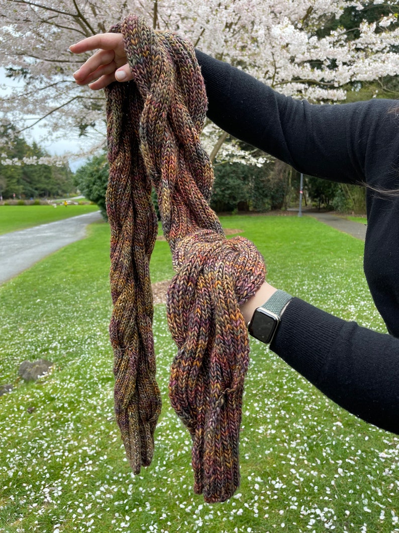 May include: A hand-knitted scarf featuring a thick, braided cable pattern. The yarn is a variegated blend of earthy tones, including deep brown, forest green, burnt orange, and muted purple, creating a textured, multi-colored appearance.