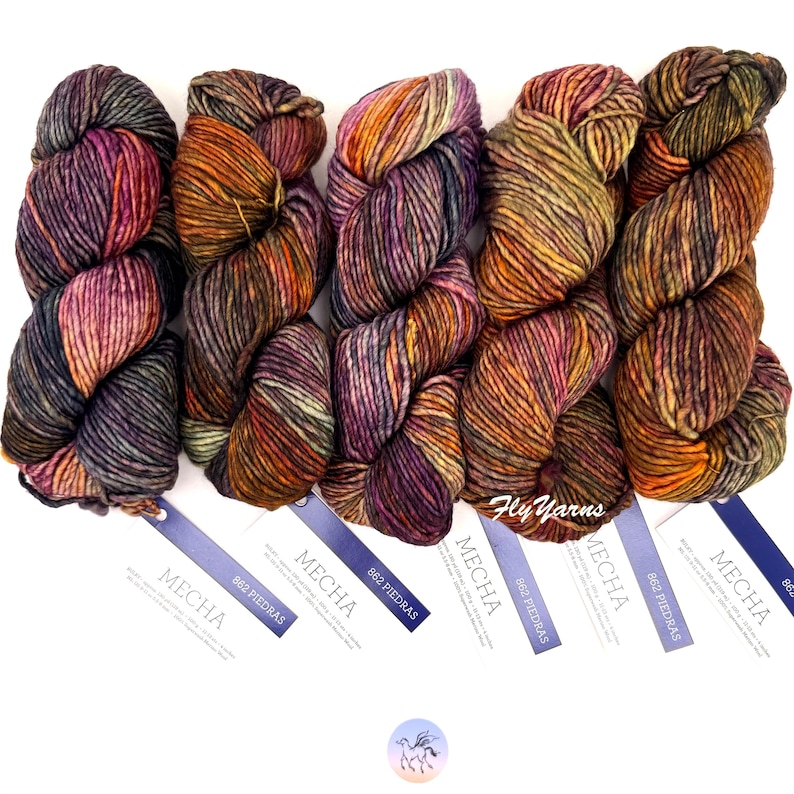 May include: Five skeins of Mecha 862 Piedras yarn arranged side by side. Each skein features a variegated blend of purple, orange, yellow, and dark green tones. Attached to each skein is a white rectangular tag with the text Mecha 862 Piedras.