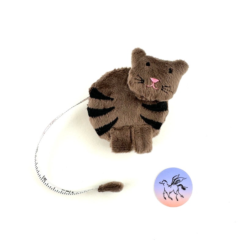May include: A brown and black striped plush cat-shaped tape measure with a pink nose and whiskers. The tape measure is curled up and the tail is extended. A small white circle with a black silhouette of a winged horse is in the background.