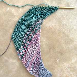 May include: A crescent-shaped knitting project in progress on a single gold-toned needle. The piece features a garter stitch texture transitioning from deep forest green to variegated sections of dusty pink, mauve, and grey with openwork lace.
