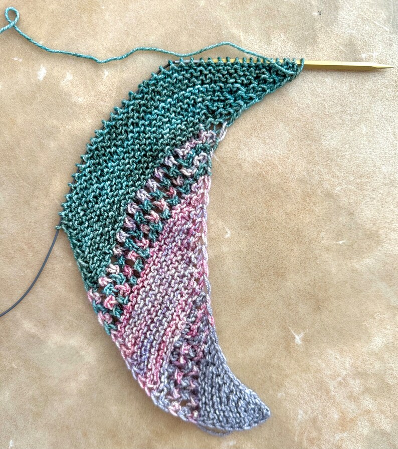 May include: A crescent-shaped knitting project in progress on a single gold-toned needle. The piece features a garter stitch texture transitioning from deep forest green to variegated sections of dusty pink, mauve, and grey with openwork lace.