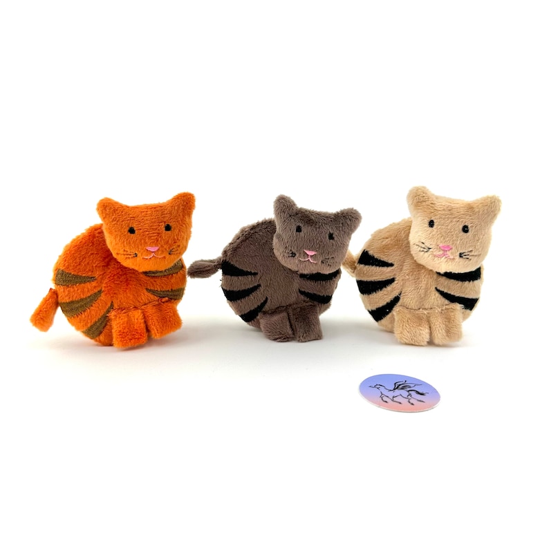 May include: Three plush toy cats in different colors: orange, gray, and beige. Each cat has black stripes and pink embroidered noses.