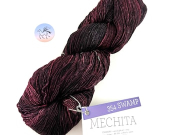 Puede incluir: Una madeja de hilo de peso fingering de color morado oscuro y negro llamada "Mechita" en el color "354 Swamp". El hilo es 100% lana merino superwash y tiene un metraje de 430 yardas (391 metros) por madeja de 100g.
