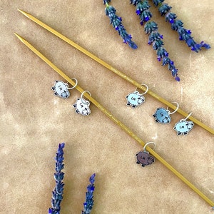 May include: Two gold-colored knitting needles with sheep-shaped stitch markers. The needles are labeled "Kollage Square Needles US 7/4.5 mm 10" (25 cm)". Lavender sprigs with purple flowers are arranged around the needles, creating a craft-themed image.