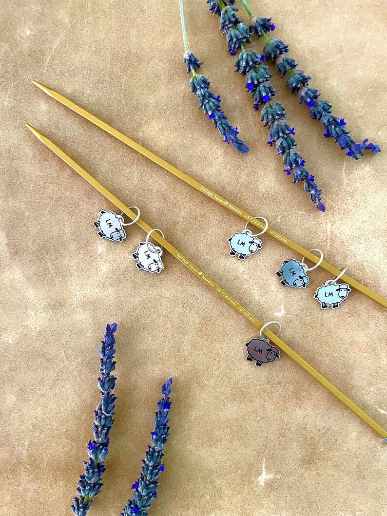 May include: Two gold-colored knitting needles with sheep-shaped stitch markers. The needles are labeled "Kollage Square Needles US 7/4.5 mm 10" (25 cm)". Lavender sprigs with purple flowers are arranged around the needles, creating a craft-themed image.