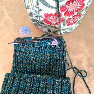 Pu&ograve; includere: Un cappello lavorato a maglia in corso d'opera, con filato verde acqua, blu e verde. Ferri da maglia neri e un segnapunti rosa sono in posizione. &Egrave; visibile una piccola spilla con un disegno di cavallo e una borsa floreale.
