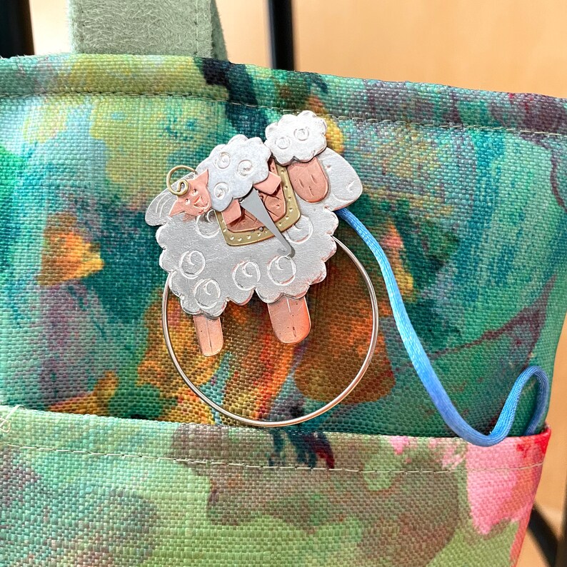 Pode incluir: Um amuleto de ovelha prateado e cobre com um anel preso. O amuleto est&aacute; pendurado em um cord&atilde;o azul em uma bolsa de tecido floral verde e branca.