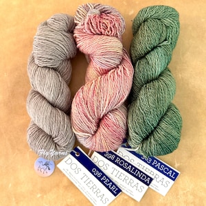 May include: Three skeins of Dos Tierras yarn in grey, variegated pink and orange, and green. Each skein has a white tag with blue text reading Dos Tierras, with colour names Pearl, Rosalinda, and Pascal. A small round tag features a horse logo.