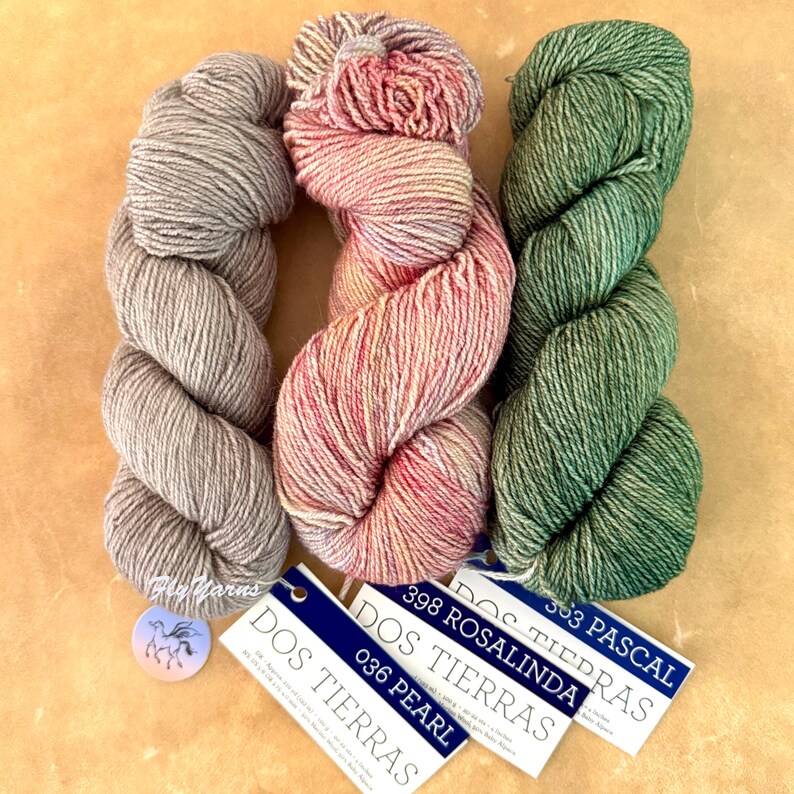May include: Three skeins of Dos Tierras yarn in grey, variegated pink and orange, and green. Each skein has a white tag with blue text reading Dos Tierras, with colour names Pearl, Rosalinda, and Pascal. A small round tag features a horse logo.