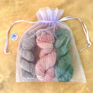 May include: A sheer lavender organza drawstring bag containing three skeins of yarn in grey, variegated pink, and green. A small circular white tag with a blue winged horse logo and the text Fly Yarns is positioned to the left of the bag.