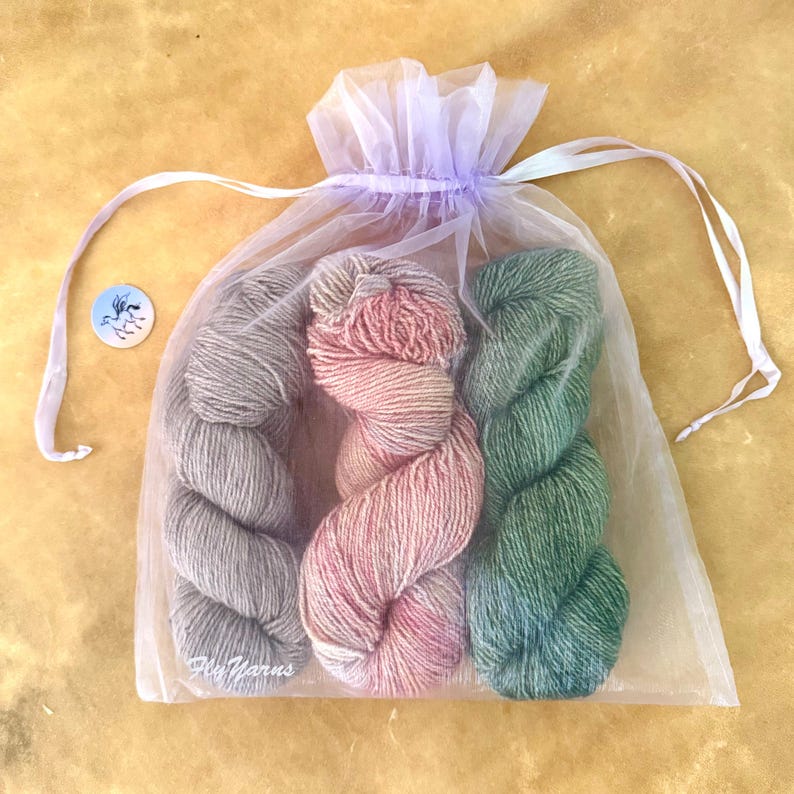 May include: A sheer lavender organza drawstring bag containing three skeins of yarn in grey, variegated pink, and green. A small circular white tag with a blue winged horse logo and the text Fly Yarns is positioned to the left of the bag.