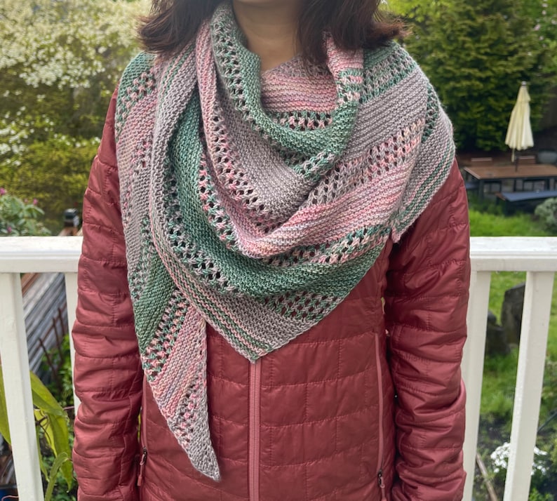 May include: A triangular knitted shawl featuring a variegated colour palette of sage green, soft pink, and muted grey. The accessory displays a combination of garter stitch sections and decorative eyelet lace rows, creating a textured, openwork pattern.