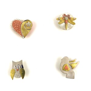 Pode incluir: Quatro alfinetes de metal com diferentes designs: um cora&ccedil;&atilde;o com uma estrela, uma lib&eacute;lula, uma coruja e um p&aacute;ssaro com um cora&ccedil;&atilde;o. Os alfinetes s&atilde;o feitos de prata, ouro e cobre.