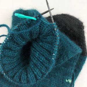 May include: Close-up of a teal and black knitted hat with a knitting needle and stitch marker. The hat is being worked in the round and the needle is holding a loop of yarn.