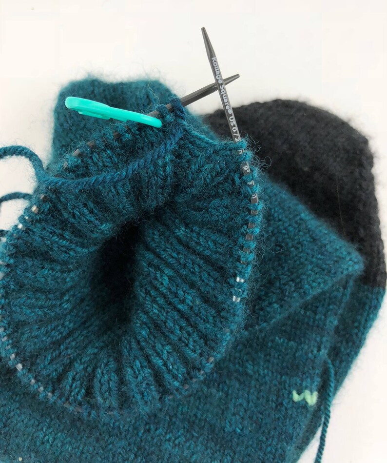 May include: Close-up of a teal and black knitted hat with a knitting needle and stitch marker. The hat is being worked in the round and the needle is holding a loop of yarn.