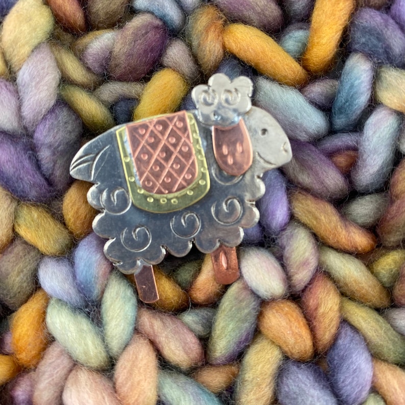 Pode incluir: Um broche de metal prateado em forma de ovelha com uma sela estampada em cobre e verde. A ovelha est&aacute; em p&eacute; sobre um fundo de fio multicolorido.