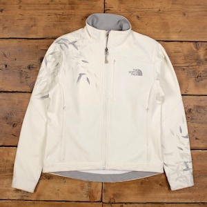 May include: Cream-colored The North Face jacket with a full zipper and a gray collar. The jacket features a leaf pattern in gray on the shoulders and sleeves. The North Face logo is on the chest.