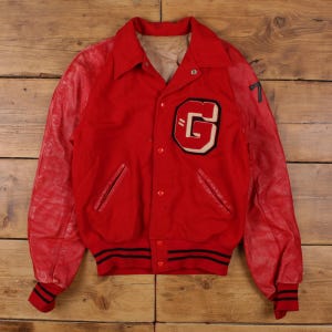 Könnte beinhalten: Vintage rote Letterman-Jacke mit Lederärmeln. Die Jacke hat einen großen weißen und schwarzen "G"-Aufnäher auf der Brust und die Nummer "7" auf dem rechten Ärmel. Schwarz-weiße Streifen akzentuieren die Manschetten und den Bund.