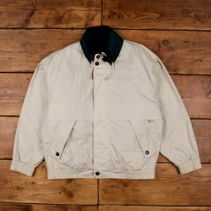 May include: A beige windbreaker jacket featuring a dark green collar, a front zipper closure with a buttoned storm flap, two side pockets with buttoned flaps, and elasticized cuffs and hem. The jacket includes a drawstring at the neckline.