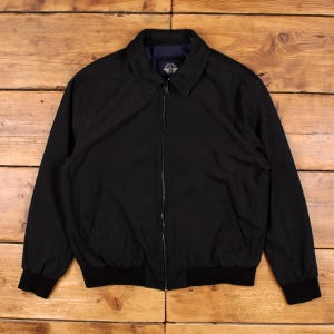 May include: A black zip-up jacket featuring a classic collar, long sleeves with ribbed cuffs, and a ribbed hem. The garment includes a central metal zipper closure and two front pockets. A small brand label is visible on the inner neck area.