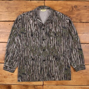 Peut inclure: Chemise boutonnée de camouflage avec un motif de rayures verticales marron et grises et des accents de feuilles vertes. La chemise a quatre poches avant et des boutons noirs. Le col est de style classique.