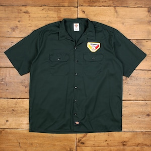 May include: Dark green short-sleeved work shirt with two chest pockets and a button-down front. The shirt features a patch on the upper left chest with text and a logo. The shirt is made of a durable material.