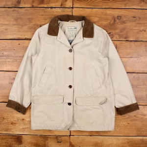 May include: A tan jacket with a brown corduroy collar and cuffs. The jacket has a button-up front, two front pockets, and a small logo on the collar. The jacket is laid flat on a wooden surface.