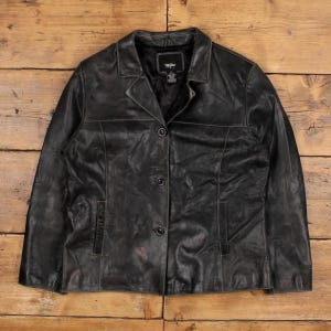 May include: A black leather jacket with a classic design. The jacket features a collared neckline, three buttons, and two front pockets. The leather has a slightly distressed appearance, giving it a vintage look. The jacket is laid flat on a wooden surface.