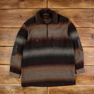 Puede incluir: Un jersey de lana vintage con un estampado de rayas marrones, grises y beige. La chaqueta presenta un cuello, un cierre de cremallera de cuarto, dos bolsillos delanteros y coderas de cuero. La etiqueta dice "Nontak".