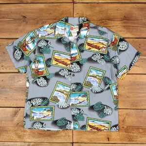 May include: A short-sleeved, button-down shirt with a light gray background. The shirt features a repeating pattern of framed beach scenes with palm trees, boats, and figures, along with green and white tropical leaves. The shirt has a classic collar.