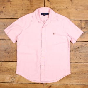 May include: A pale pink short-sleeved shirt with a button-down front. The shirt features a classic collar and a small embroidered logo on the chest. The fabric appears lightweight. The shirt is displayed on a wooden surface.