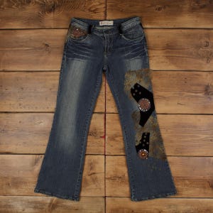 May include: Blue denim jeans with decorative patches and embellishments. The jeans feature a flared leg, a faded wash, and decorative beading around the pockets and on the patches. The patches are a mix of textures and colors.