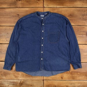 May include: A long-sleeved denim shirt in a medium blue shade, with a mandarin collar and a single chest pocket. The shirt has a button-down front with white buttons and a relaxed fit, suitable for casual wear.