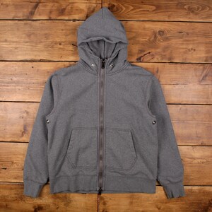 Könnte beinhalten: Ein grauer Kapuzenpullover mit Reißverschluss, Kapuze und zwei Vordertaschen. Die Jacke hat einen durchgehenden Reißverschluss und eine Kapuze mit Kordelzug. Der Stoff scheint ein weiches Material mittlerer Stärke zu sein. Der Hoodie liegt flach auf einer Holzoberfläche.