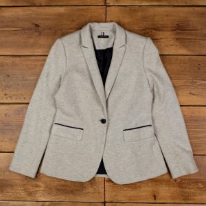 May include: A light grey heathered blazer featuring a single dark button closure, notched lapels, and two front pockets with dark trim. The interior lining is black, and a small brand tag is visible at the centre of the inner neckline.