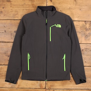 May include: A dark grey jacket with a black collar and neon green zip detailing. The North Face logo is on the chest. The jacket features two side pockets and a full-length zip.