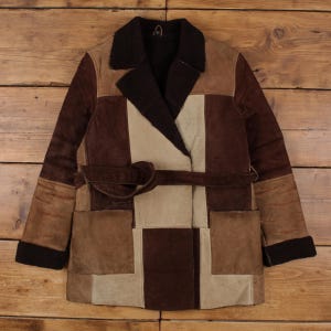 May include: A patchwork suede coat in shades of brown and beige. The coat features a wide collar, a belt, and two front pockets. The sleeves and collar are trimmed with a dark brown, fuzzy material. The coat is laid flat on a wooden surface.
