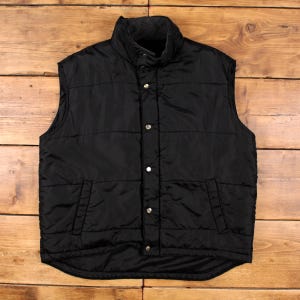 Puede incluir: Un chaleco negro acolchado con cuello alto y cierre de botones. El chaleco tiene dos bolsillos delanteros y un forro polar gris alrededor del cuello. El chaleco está hecho de un material brillante y resistente al agua.