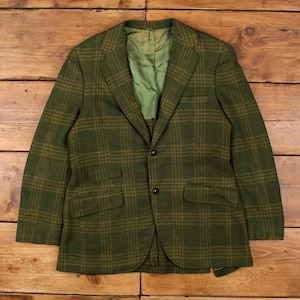 May include: Green and brown plaid blazer with two front pockets and black buttons. The blazer has a classic design and a green lining. The fabric has a textured appearance. The blazer is laid flat on a wooden surface.