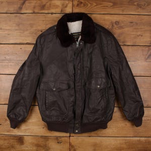 Peut inclure: Veste de bombardier en cuir marron avec un col en fausse fourrure marron foncé et une doublure intérieure blanche. La veste a deux poches avant avec fermetures à boutons et un ourlet et des poignets côtelés. La veste est présentée sur une surface en bois.