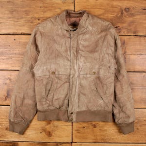 Könnte beinhalten: Hellbraune Wildleder-Bomberjacke mit geripptem Kragen, Manschetten und Saum. Die Jacke hat zwei Vordertaschen mit Knopfverschluss und eine Knopfleiste. Die Jacke ist neutralfarben und hat einen klassischen Vintage-Stil.