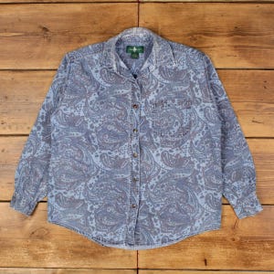 Può includere: Una camicia a maniche lunghe abbottonata in una tonalità blu sbiadito con un motivo paisley blu scuro su tutta la superficie. La camicia presenta un colletto a punta, una tasca sul petto a sinistra e un'abbottonatura frontale con bottoni marroni.