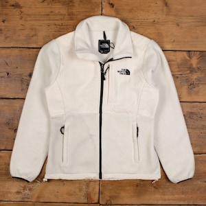 May include: Cream fleece jacket with a black zip and two side pockets. The jacket has the brand logo on the chest and a small tag near the collar. The jacket is made of a soft, textured material. A classic outdoor garment.