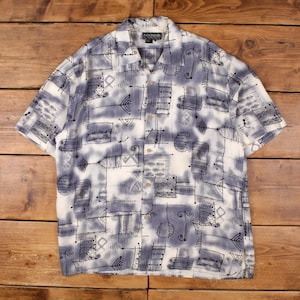May include: Short-sleeved button-down shirt with a white base and a blue and black abstract pattern. The shirt features a collared neckline and wooden buttons. The design includes geometric shapes and lines.